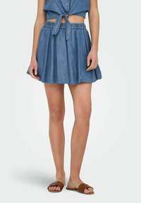Jeans krilo z elastičnim pasom, razširjenega kroja in svetlo modre barve. V kombinaciji z rjavimi pletenimi sandali, kar povečuje sproščen videz.