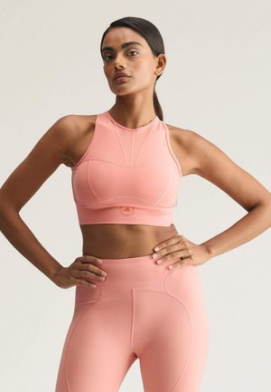 Kvinde står med hænderne på hofterne iført matchende pink sports-bh og høj talje leggings mod ensartet baggrund.