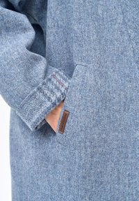Zbliżenie na dłoń w kieszeni niebieskiego, teksturowanego, wełnianego płaszcza z podwiniętym mankietem i małą skórzaną metką z logo.