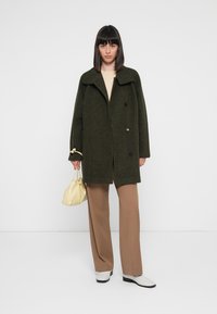 Olive green wool coat with a notched collar and double-breasted buttons, worn over beige top and brown trousers. White ankle boots and yellow bag.