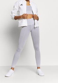 Setet inkluderar en vit zipped jacka och en matchande lång ribbad crop top och leggings, allt i ljusgrått med ett strukturerat mönster. Vita sneakers synliga.