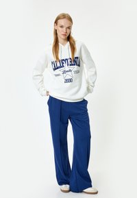 Weißer Kapuzenpullover mit blauer Schrift und Stern-Grafik, kombiniert mit blauen weitem Hosen. Das Model steht vor einem schlichten Hintergrund.