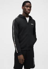 Μαύρο φούτερ με φερμουάρ και κουκούλα. Διαθέτει αντίθετες λευκές ρίγες κατά μήκος των μανικιών. Το υλικό έχει λεία, απαλή υφή. Συνδυάζεται με μαύρο σορτς.