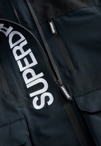 Giacca nera in nylon con un prominente logo bianco ricamato. Tasche con cerniera e tiranti neri, tessuto liscio su tutta la superficie.