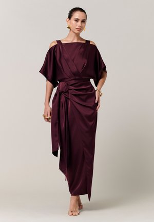 Frau in einem ärmellosen, asymmetrischen, tief violetten Satinkleid mit Wickeldetail, goldenen Ohrringen, Armband und offenen Absätzen, die nach vorne geht.