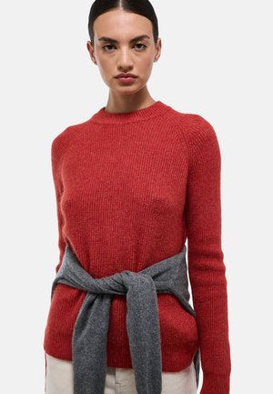 Pull en tricot rouge avec un col rond et une texture côtelée. Une ceinture cravate grise est nouée à la taille, ajoutant un contraste au look.