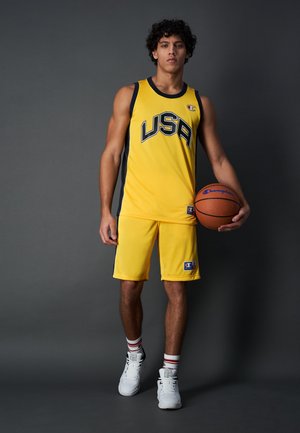 Camiseta y pantalones cortos de baloncesto amarillos con "USA" en texto negro. Presenta un logo en el pecho y en el costado, combinado con zapatillas blancas y calcetas a rayas.