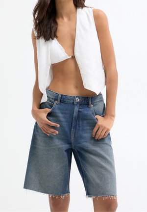Kvinde iført en hvid ærmeløs vest, hvidt blonde-bikinitop og løstsiddende, knælange denimshorts med hænderne i lommerne.