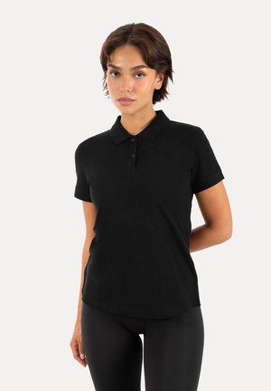 Frau mit kurzen braunen Haaren, die ein schwarzes kurzärmeliges Poloshirt und schwarze Hose trägt, steht vor einem einfarbigen hellen Hintergrund.