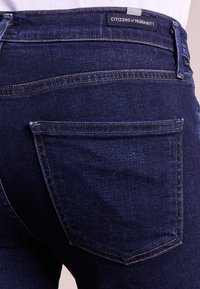 Mörkblå denimjeans med en bakficka, sömdetaljer och en etikett med "CITIZENS of HUMANITY" ovanför midjan.