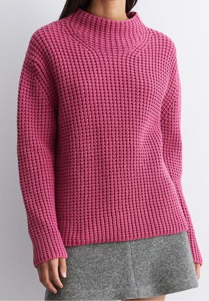 Femme portant un pull en maille rose texturé à manches longues et col montant, associé à une jupe en laine grise.