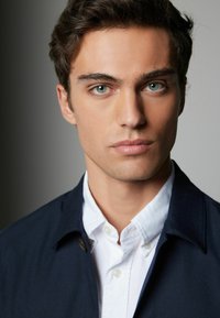 Portrait of a person with tousled dark hair, light skin, wearing a navy coat over a white button-up shirt, focusing on facial features and expression.