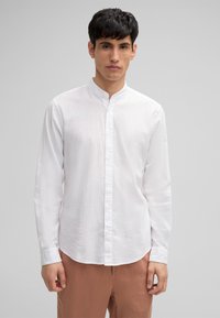 Camicia bianca a maniche lunghe con colletto alla cinese e chiusura a bottoni, caratterizzata da un tessuto texturizzato. Abbinata a pantaloni color marrone chiaro.