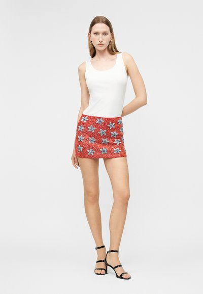 Γυναίκα που στέκεται με το χέρι πίσω από την πλάτη, φορώντας λευκό αμάνικο τοπ, κόκκινη μίνι φούστα με μπλε σχέδια αστεριών και μαύρα σανδάλια με τακούνι.