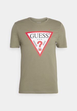 Olivenfarvet bomulds t-shirt med korte ærmer, der har et trekantet logo design i rød og sort, med påskriften "GUESS U.S.A. WASHED JEANS."
