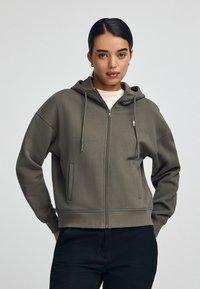Felpa con cappuccio e zip in verde oliva, realizzata in tessuto morbido. Presenta tasche frontali, cordini e un piccolo logo sul petto.