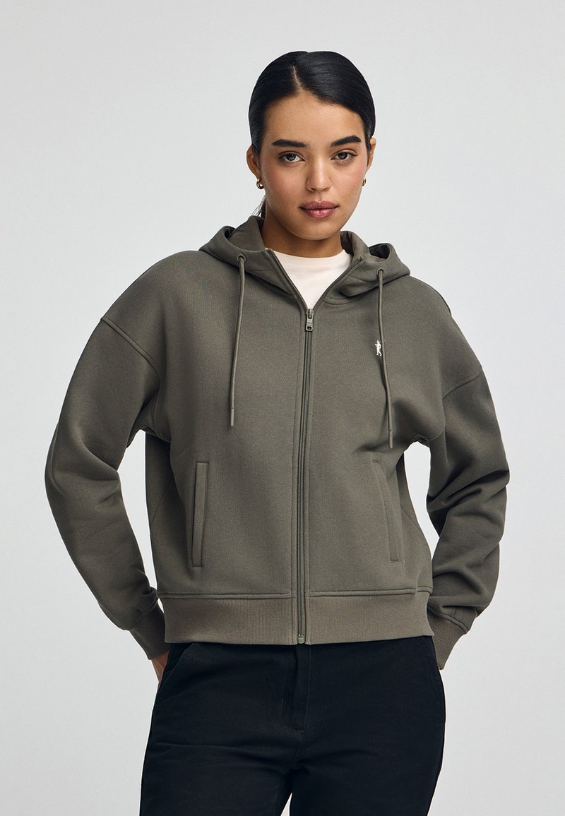Felpa con cappuccio e zip in verde oliva, realizzata in tessuto morbido. Presenta tasche frontali, cordini e un piccolo logo sul petto.