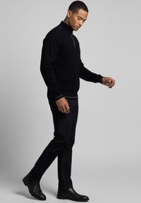 Suéter negro acanalado con cuello de cremallera hasta el cuarto y detalles en los hombros, combinado con jeans oscuros y botines negros.