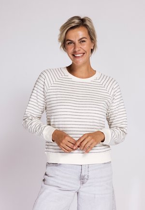 Frau lächelt, trägt ein weißes Langarmshirt mit dünnen grauen horizontalen Streifen und hellen Jeans, steht vor einem schlichten Hintergrund.