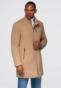 Beige Wollmantel mit hohem Kragen, ausgestattet mit Reißverschlusstaschen und einem strukturierten Design. Getragen über einem hellen Rollkragenpullover und blauen Jeans.