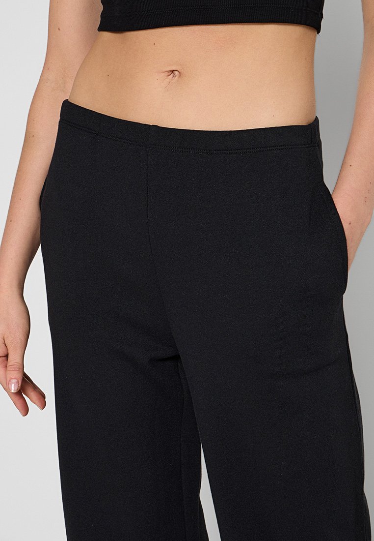 Mujer vestida con pantalones de tiro alto negros y un top corto negro, con una mano relajada y la otra en el bolsillo.