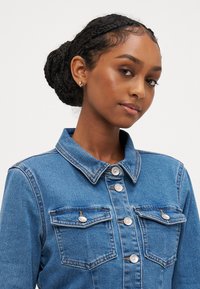 Denim jakna v srednji modri barvi s ovratnikom, dvema žepoma na prsih, srebrnimi gumbi in kontrastno šivano obrobo. Gladka tekstura.