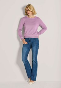Frau mit kurzem blonden Haar, die einen lavendelfarbenen Pullover, blaue Jeans und weiße Absätze trägt, posiert vor einer schlichten hellen Wand.