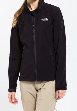 Person wearing a black zip-up North Face jacket with two side pockets and beige pants against a plain white background.