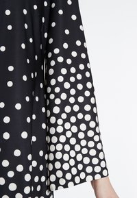 Schwarzer Stoff mit großen weißen Punkte, lässiger Schnitt und weite Ärmel. Glatte Textur mit dezentem Glanz.