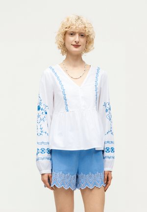 Ξανθό άτομο φοράει λευκό μπλουζάκι με μπλε κέντημα και ανοιχτό μπλε σορτς με κυματιστό δαντελένιο τελείωμα, στέκεται μπροστά σε μονόχρωμο φόντο.