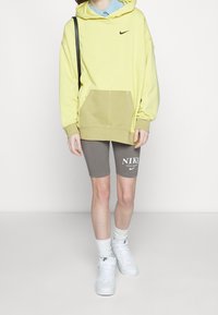 Felpa oversized gialla Nike con tasca a marsupio, abbinata a pantaloni corti da bicicletta grigi con logo bianco, e sneakers bianche con suola testurizzata.