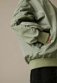 Olive zelena bomber jakna z gladkim, sijočim materialom, elastičnimi manšetami in nastavljivim pasom z subtilnimi teksturami.