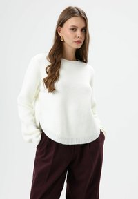 Pull en tricot blanc avec un col rond, des poignets côtelés et des détails de boutons sur le côté, associé à un pantalon taille haute bordeaux foncé.