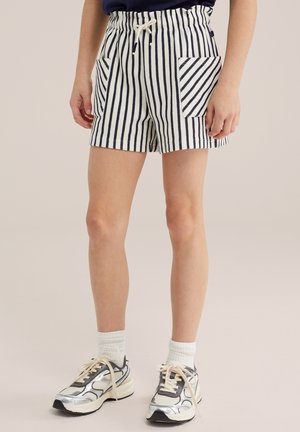 Osoba nosiaca bielo-námornícko modré pruhované kraťasy s prednými vreckami, biele ponožky a strieborno-biele tenisky, stojaca na béžovej podlahe.