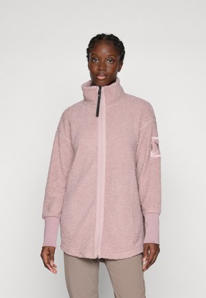Frau, die eine hellrosa Zip-Jacke aus Fleece mit hohem Kragen und beigen Hosen trägt, vor einem schlichten hellen Hintergrund steht.