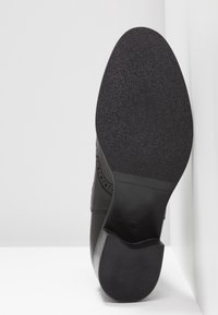 Chaussure en cuir noir avec une semelle en caoutchouc texturée et des perforations décoratives sur le côté. Dotée d'un talon bas et de lignes épurées.