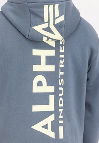 Siva dokolenka iz mešanice bombaža, ki ima na hrbtni strani odločno belo besedilo "ALPHA INDUSTRIES" v navpični orientaciji in kapuco s vrvico.