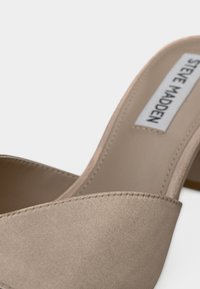 Sandale à talon haut en satin beige avec bout ouvert, forme courbée et design minimaliste. Comprend une étiquette "STEVE MADDEN" sur la semelle intérieure.
