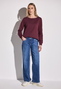 Felpa bordeaux con scollo rotondo, abbinata a jeans a gamba larga blu chiaro. L'outfit è completato da sneakers bianche e dettagli minimi.