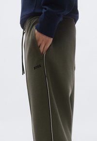 Olívazöld futónadrág puha anyaggal, oldalzsebekkel, hímzett "BOSS" logóval és kontrasztos fehér szegéllyel az oldalakon.