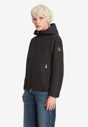 Giovane con capelli biondi corti che indossa una giacca nera con cappuccio e jeans blu, in piedi davanti a uno sfondo semplice.