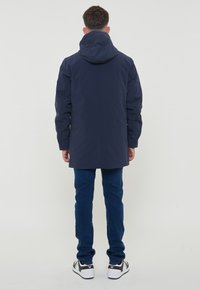 Parka navy con cappuccio, caratterizzato da una vestibilità dritta e maniche lunghe, abbinato a jeans blu scuro e sneakers bianco e nere.