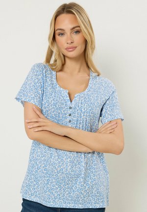 Blonde Frau mit blauen Augen trägt eine hellblaue gemusterte Bluse mit kurzen Ärmeln und Knöpfen, verschränkt die Arme vor einem einfarbigen Hintergrund.