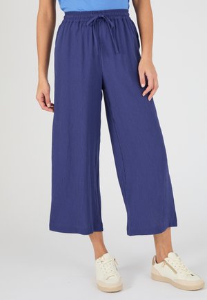 Persona che indossa pantaloni blu navy larghi e corti, con vita elastica e coulisse, abbinati a sneaker bianche casual.