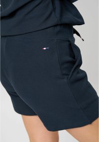Námořnické modré sportovní šortky s texturovanou látkou, elastickým páskem, bočními kapsami a malým detailem loga v červené, bílé a modré barvě.