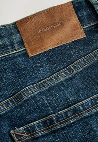 Sinised denim teksad, millel on tume pesu, pruun nahklabel embosseeritud tekstiga ning nähtavad õmblused taskute ja õmbluste ääres.