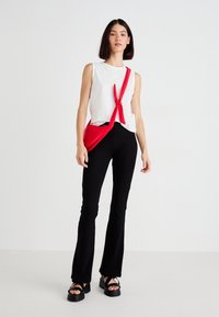 Top bianco senza maniche legato in vita, pantaloni neri a palazzo e borsa a tracolla rossa. Sandali neri spessi con fasce larghe. Sfondo neutro.