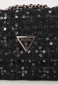 Borsa a tracolla nera con paillettes e superficie testurizzata, tracolla in metallo dorato e logo triangolare GUESS sul fronte.