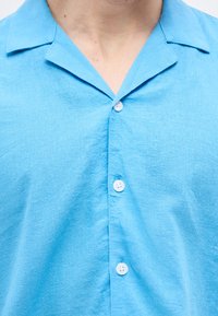 Ανοιχτό μπλε λινό πουκάμισο με ανοιχτό γιακά και τέσσερα λευκά κουμπιά κλεισμένα, αποκαλύπτοντας τον λαιμό και το πάνω μέρος του στήθους του φορέα.