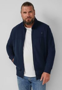 Felpa zip-up blu marina con colletto alto, in tessuto morbido e con un interno bianco a contrasto, abbinata a jeans azzurri chiari. Design semplice.